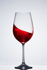 Wave of the red wine in the pure fragile wineglass standing against light background with reflection.