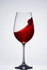 Splashing and moving red wine in the pure wineglass standing against light background with reflection on the stand.
