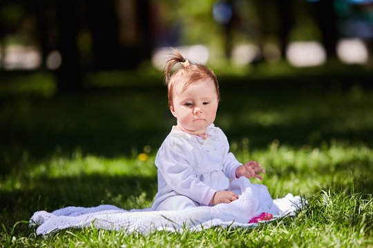 Cute baby girl sitting on the green grass in the city park at warm summer day.