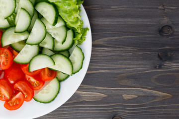 Salad of fresh cherry tomatoes, cucumber and lettuce leaves on a white plate on a dark wooden table. View from above. Place for text.