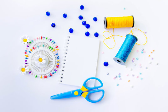 Sewing spools of blue and yellow caprone threads, blank spiral notepad, plastic scissors, glass beds and colorful pins on white background