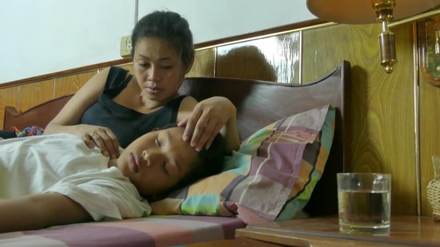 Medium bedside view of a multicultural Asian mother with her sick son. She reaches to the bedstand to give him a glass of water, he drinks it. One of a series