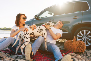 Portrait of happy young adult couple with dog on roadtrip. Man sitting on plaid with woman. Outdoor...