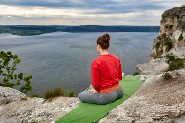 Naklejka na ściany i meble Rear view of young woman sitting in lotus position on the rock over the river.