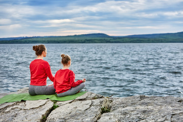 Fototapeta premium Rear view of mother and daughter practicing yoga on rocks near river.