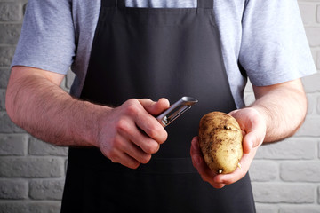 man in black apron holding potatoes and knife for peeling in his hands, on background of brick wall