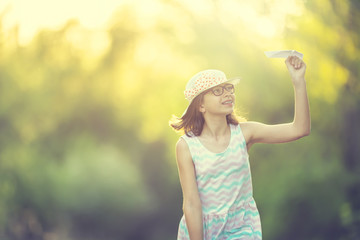 Cheerful pre-teen girl playing with paper plane on the park at sunrise. Girl with glasses and teeth...