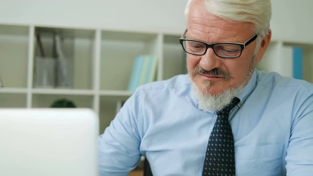 Serious midle aged man in glasses typing on his laptop. Office background. Caucasian man concentrated working in office on laptop. Close up. Indoor.