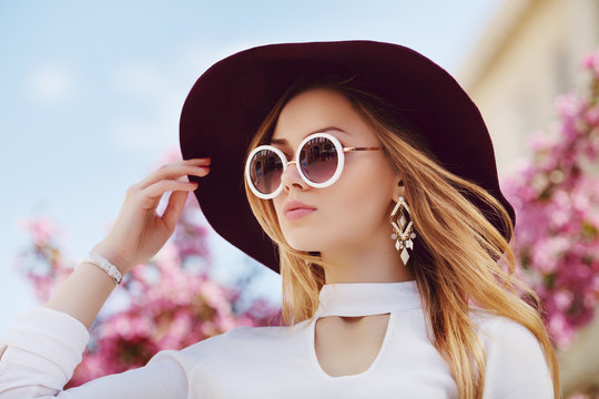 Outdoor close up portrait of young beautiful girl posing in street, near blooming tree. Model wearing stylish round sunglasses, wide-brimmed hat, white shirt. City lifestyle. Female fashion concept. 