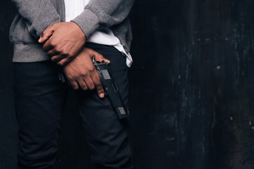 Unrecognizable armed black criminal man studio shoot. Gangster guy with gun in hand on dark...