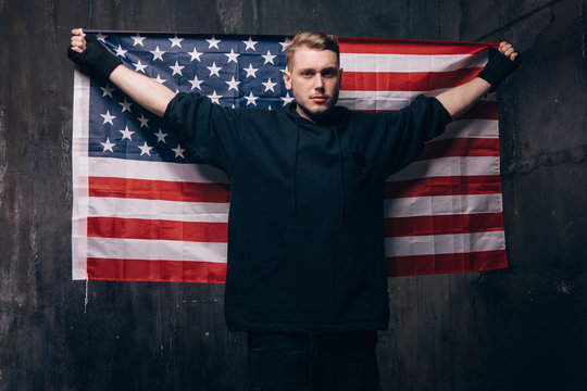 Tired USA patriot holds national flag. Strong man in black cloth on dark background. Independence day, confidence, pride, fidelity to the nation, memorial day concept