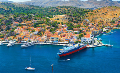 View on beautiful traditional Greek houses on Symi island green hills, yacht sea port, tourist ferryboat at Aegean Sea blue bay. Mediterranean MSC cruises. Greece islands holiday vacation tours trip