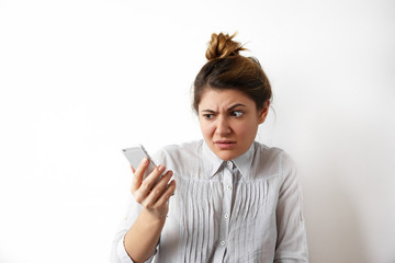 Human emotions, feelings and reactions. Attractive young woman with hair bun looking at mobile phone seeing bad news photos having scared or shocked facial expression, raising eyebrows