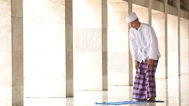  
Religious young muslim person wearing islamic clothes and praying with pose stand up from a prostration in the mosque
