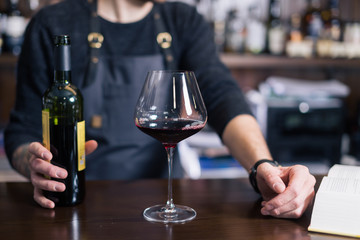 Sommelier examining wine. Cropped image of confident male sommelier examining wine while smelling it and leaning at the wooden table with wine shelf in the background