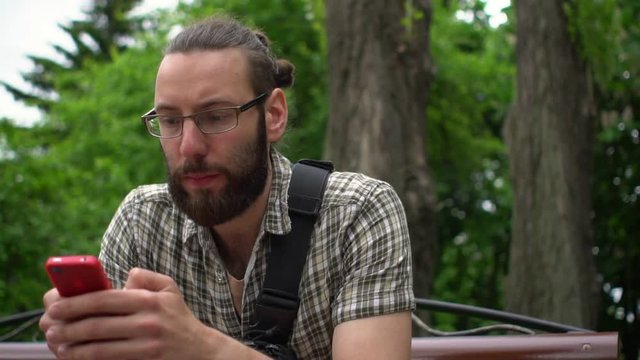 Handsome man with beard and glasses typing text in a park on his smartphone