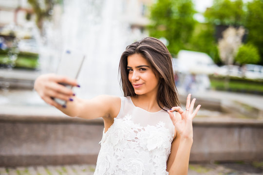 Happy young female traveler taking selfie on street with okay gesture