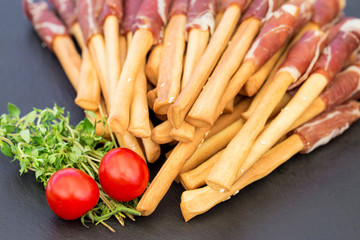 Bread sticks grissini with prosciutto ham on a black stone board. Typical italian snack .