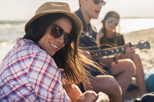 group of friends with guitar on the beach party on sunset- Young hipster people on summer vacation playing guitar near the sea. girl in hat is smiling and looking at camera