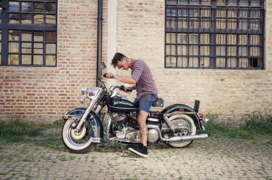 Young tattooed man sitting on a vintage motorcycle ,  side view , grungy brick wall background 