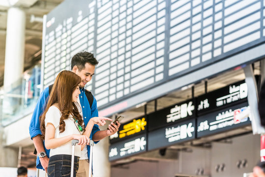 Asian couple traveler using the smart mobile phone for check-in at the flight information screen in modern an airport, travel and transportation with technology concept.