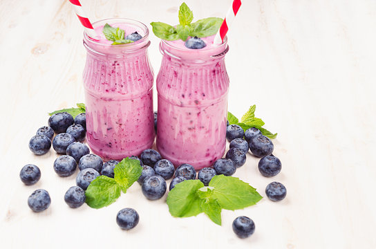Freshly blended violet blueberry fruit smoothie in glass jars with straw, mint leaves, berries. White wooden board background, copy space.