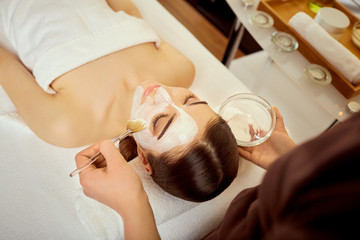 Cosmetologist puts a mask from cream on girl's face in the spa salon.