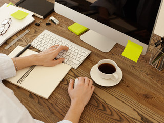 Overhead view of hipster workspace with computer PC, woman's hands on keyboard and computer mouse, cup of coffee, notepad and stationery on wooden desk. Empty space for your promotional product
