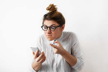 Fototapeta premium Portrait of a young indignant businesswoman with hair bun in glasses and shirt looking with anger at mobile phone. Disappointed confused female student. Negative human emotions, facial expressions