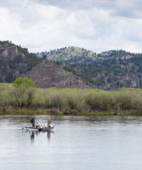 Fototapeta na wymiar Two fishermen and a third man rowing a boat in the Missouri River in Montana. Willows along the river bank and foothills are seen in the distance and cloudy sky above.