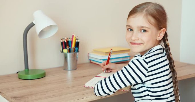 Student girl doing homework in exercise book at the table and smiling at camera