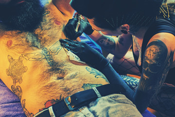 Female tattoo artist makes tattoo on bearded male torso in a salon.