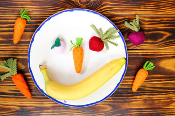 One fresh banana and fabric handmade fruits and vegetables: carrots, beet, strawberry on white ceramic plate on brown wooden background boards. Top view. Concept of creativity and healthy lifestyle