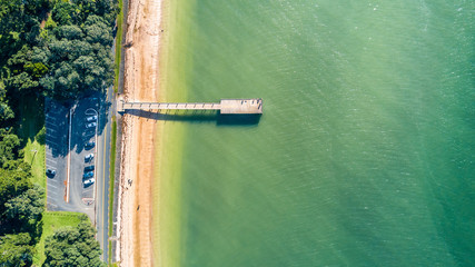 Aerial view on a sunny beach with small jetty and a car park along the road. Auckland, New Zealand