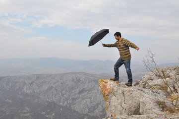 Obraz premium Young man with umbrella on a cliff edge on the top of mountain. Dangerous cliff on top of the mountain