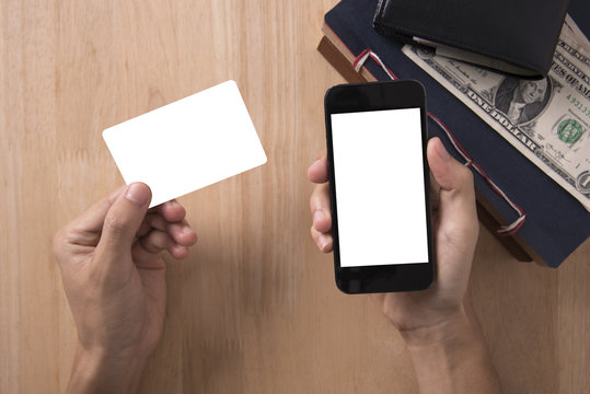Top view of hand holding credit card and smartphone with white blank screen on wood desktop.