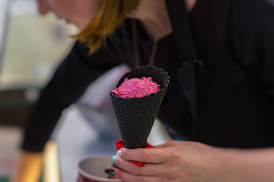 Young Saleswoman In An Ice Cream Parlor Putting Bright Pink Fruit Ice Cream Balls In Black Waffle Cone. Selective Focus, For Background Use.