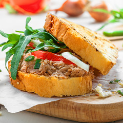 Delicious sandwich made of toasts, tuna, tomato, onion and arugula with ingredients on a white table. Traditional healthy food.
