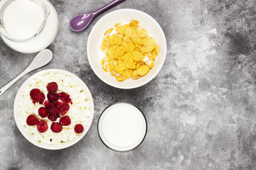 Cottage cheese in bowl with frozen raspberry and cup of cornflakes, milk in glass on a light background. Top view, copy space. Food background
