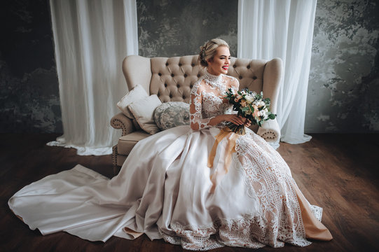 Beautiful bride in wedding lace dress with wedding bouquet with orange and white flowers. Studio, gray background, modern, sofa.