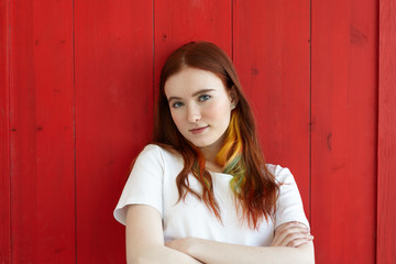 Fototapeta na wymiar Handsome girl with colored strands in ginger long hair wearing white top looking at camera with folded arms. Half body shot of green-eyed student female with crossed arms