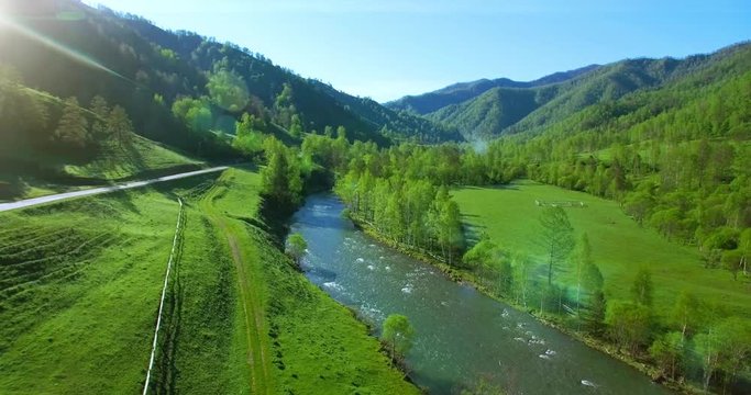 Aerial UHD 4K view. Mid-air flight over fresh mountain river and meadow at sunny summer morning. Green trees and sun rays on horizon. Rural dirt road below.