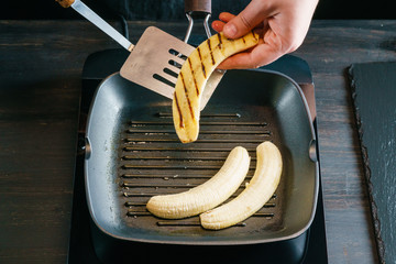 Chef's hand, taking halves of the peeled banana off the black grill-pan, with the help of a kitchen spatula. Wooden tabletop.