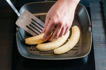 Chef's hand, taking halves of the peeled banana off the black grill-pan, with the help of a kitchen...