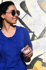 Portrait of an emotional young girl with black hair and piercings. Photo of a girl with aerosol paint cans in hands on a graffiti wall background. The concept of street art and use of aerosol paints