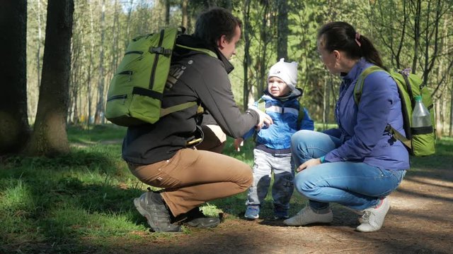 A family with a beautiful baby is going on a hike in the forest. They put on backpacks and follow the path. The sun is breaking through the leaves