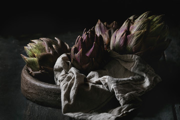 Uncooked whole organic wet purple artichokes in terracotta tray with textile over dark wooden background. Rustic style.
