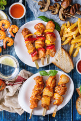 Variety of BBQ snack lunch. Plates grilled spicy prawn kebabs, chicken, pork, vegetables, mushrooms skewers, bread, french fries potatoes with sauces and greens over blue wooden background. Flat lay