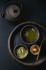 Green tea matcha powder and hot drink in black bowls standing with iron teapot, bamboo traditional tools spoon and whisk in terracotta tray over dark metal background. Top view with space