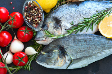 Fresh raw dorado fish with rosemary, garlic, tomatoes, pepper and lemon on a black slate background. Selective focus. Overhead view. Copy space.